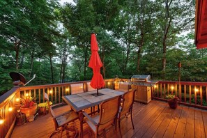 An outdoor deck with a table and chairs, a grill, and string lights. - Secluded Cherry Log Cabin - Sauna - Hot Tub - Putting Green (Blue Ridge)