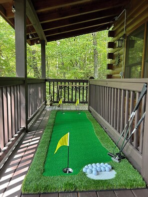 A covered deck with a miniature golf setup, including a green mat, a flag, and golf balls. - Secluded Cherry Log Cabin - Sauna - Hot Tub - Putting Green (Blue Ridge)