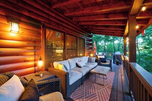 A cozy log cabin with a wooden deck, wicker furniture, and a large window overlooking a forest. - Secluded Cherry Log Cabin - Sauna - Hot Tub - Putting Green (Blue Ridge)
