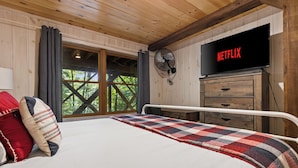 A bedroom with a bed, a TV mounted on the wall, a wooden ceiling, and a window with a view of trees.