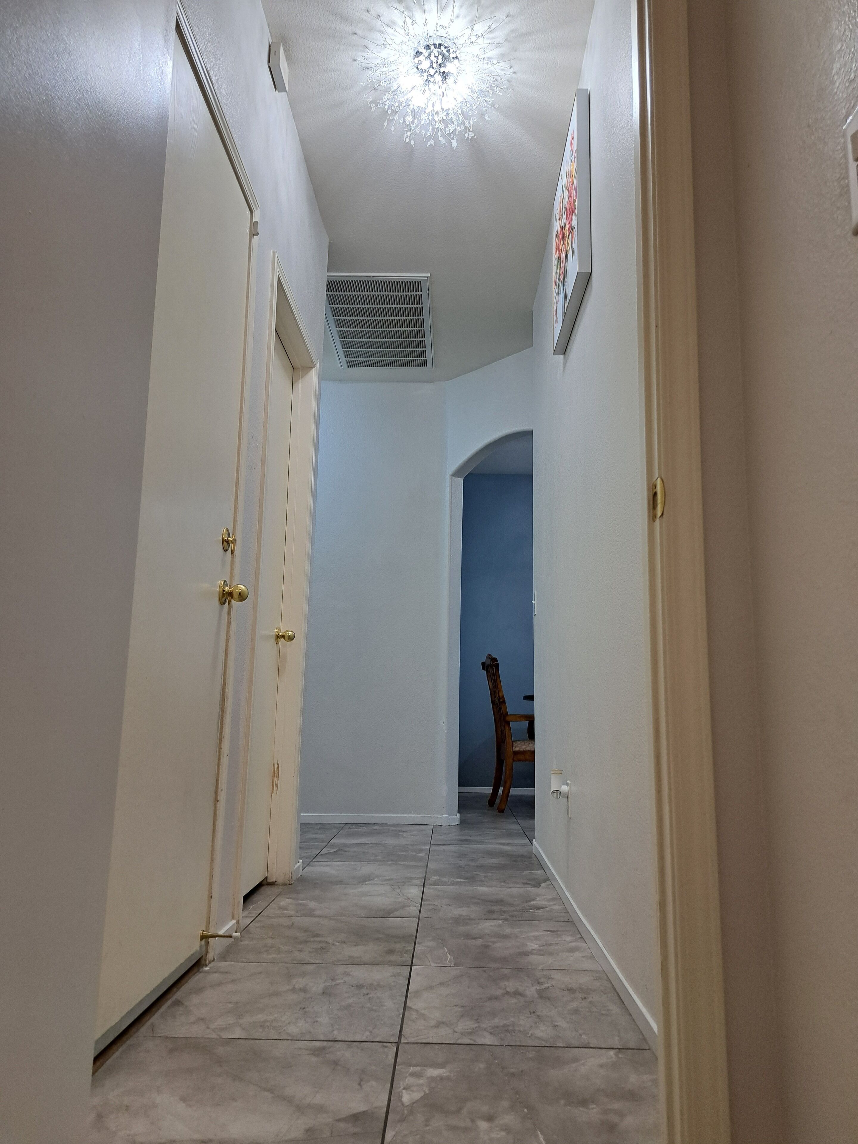 A hallway with white walls, a tiled floor, and a ceiling vent.