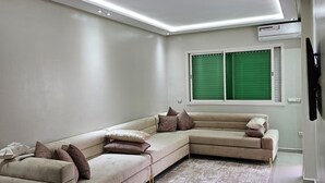 A modern living room with a L-shaped sectional sofa, recessed ceiling lighting, a patterned rug, and a window with green blinds. - Résidence Beach palace 1, apartment at the beach  (Mohammédia)