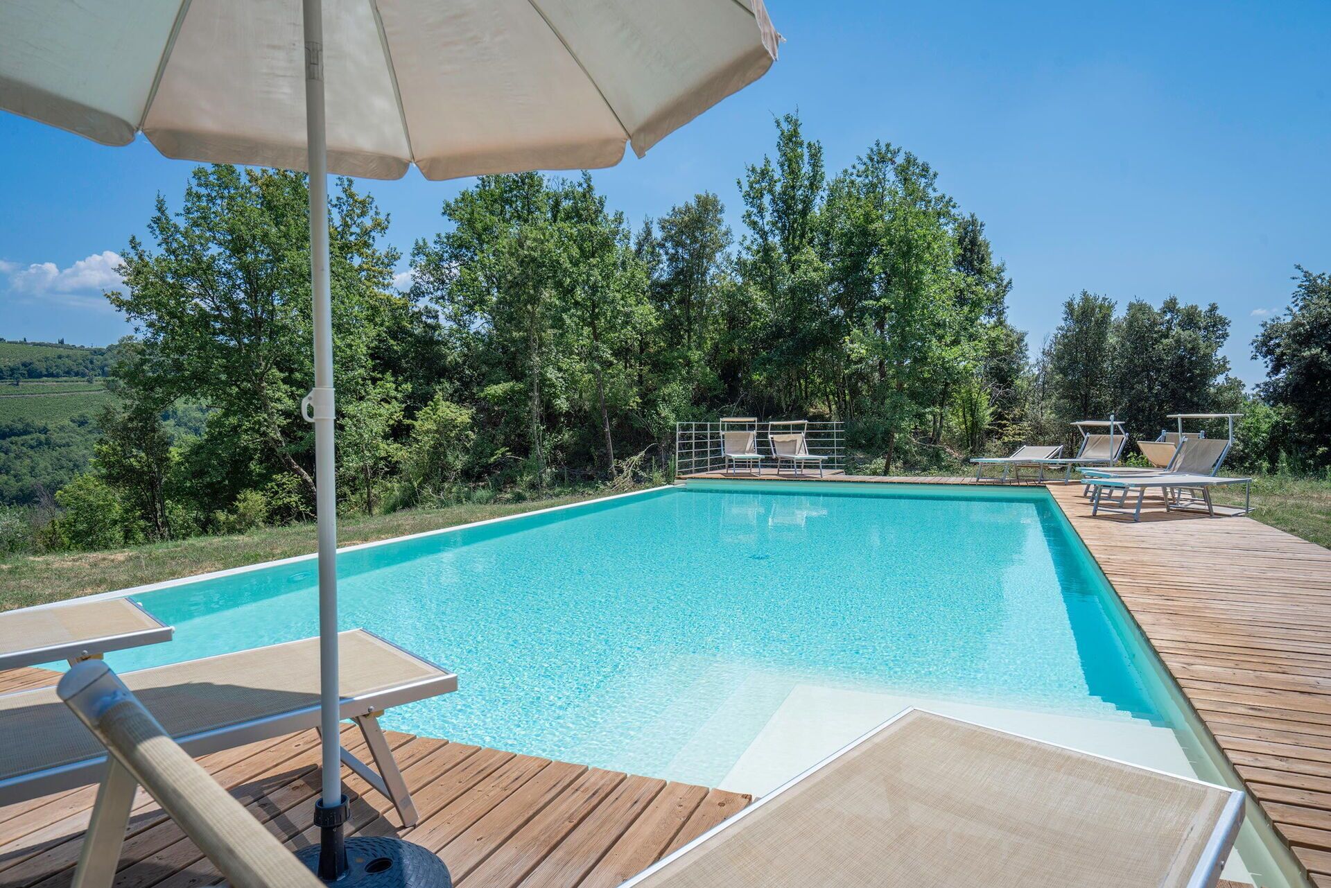 A rectangular swimming pool with clear blue water, surrounded by a wooden deck with lounge chairs and an umbrella.