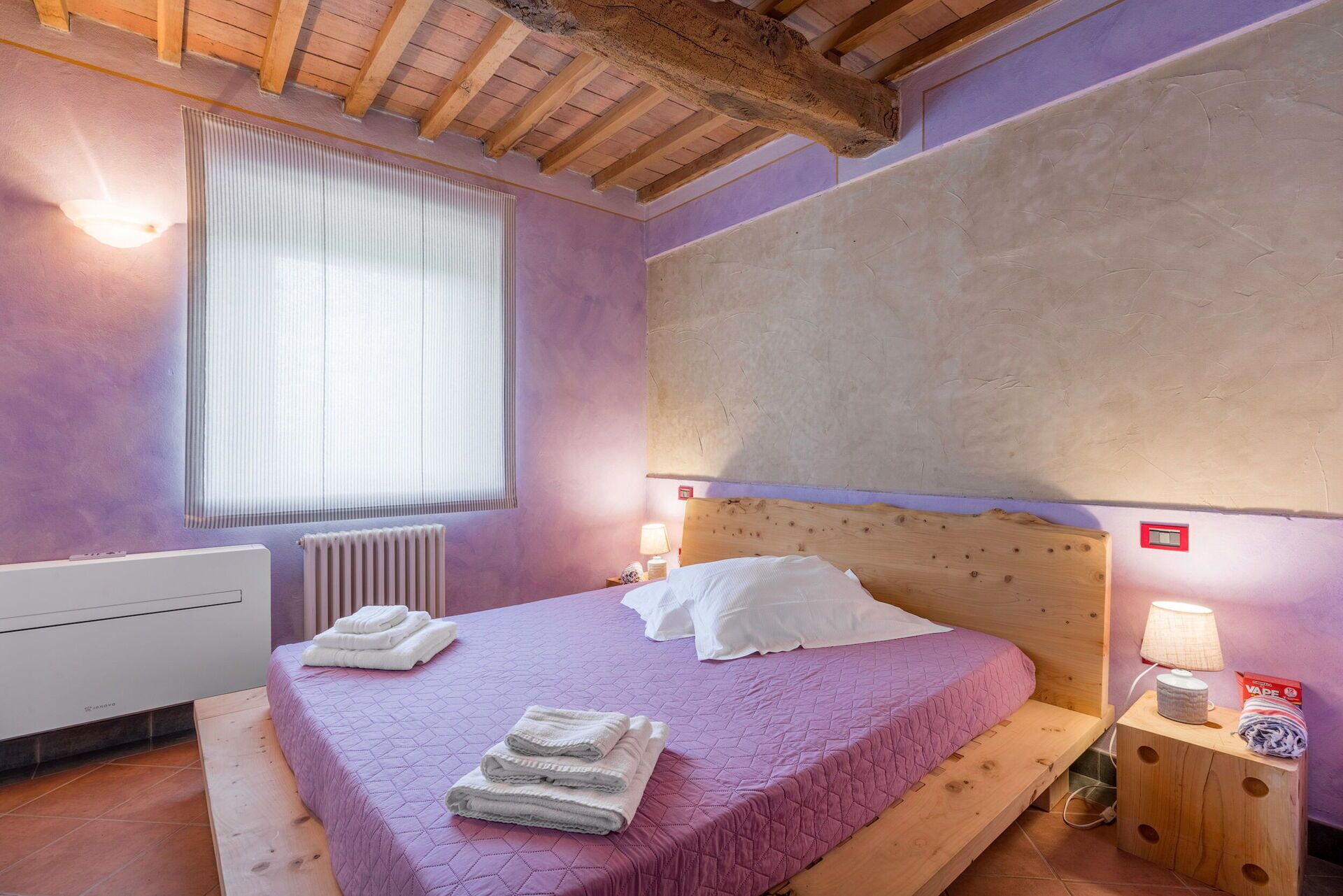 A bedroom with a wooden headboard, a large window with blinds, a radiator, and a bedside table with a lamp and a box.