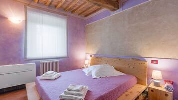 A bedroom with a wooden headboard, a large window with blinds, a radiator, and a bedside table with a lamp and a box.