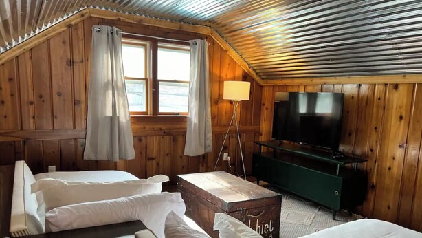 Una habitación tipo cabaña con paredes de madera, un ventanal con cortinas, una cama, una mesita de noche, una lámpara y un televisor.