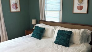 Una cama bien tendida con sábanas blancas y dos almohadas turquesa, una mesita de noche con una lámpara, y dos cuadros enmarcados en la pared.