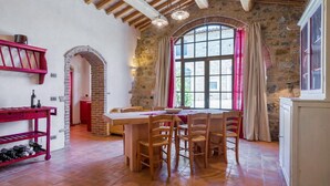 A rustic dining area with a stone wall, wooden ceiling beams, and a large arched window with pink curtains.