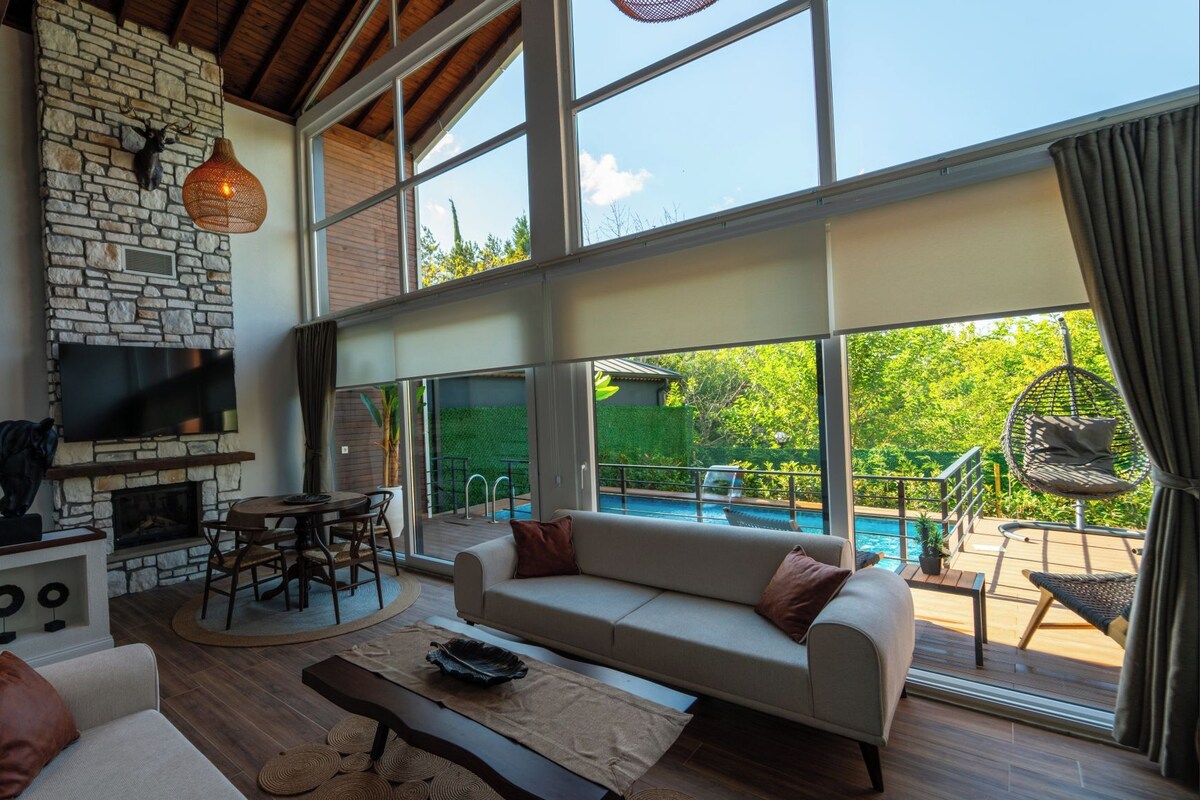 A spacious living area with a stone fireplace, a large TV, and a poolside seating area with a view of greenery.