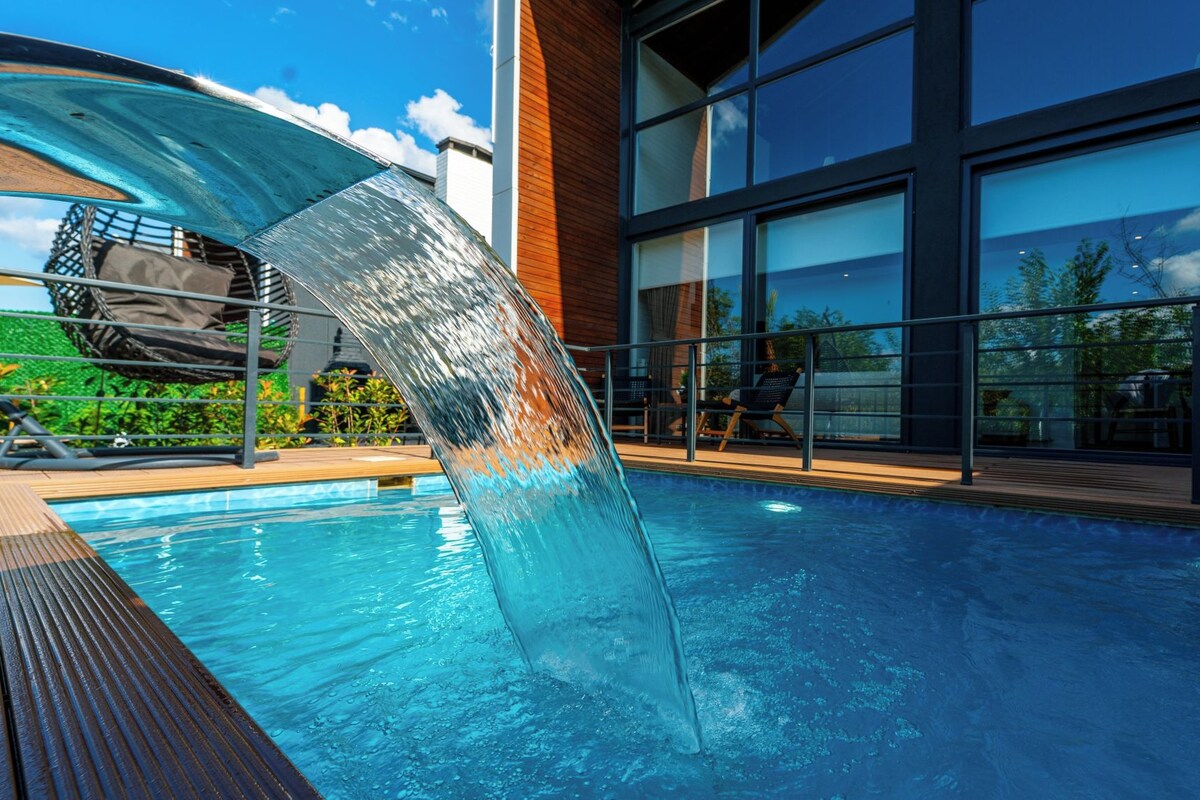 A modern pool with a clear water feature, surrounded by a wooden deck and glass railings.