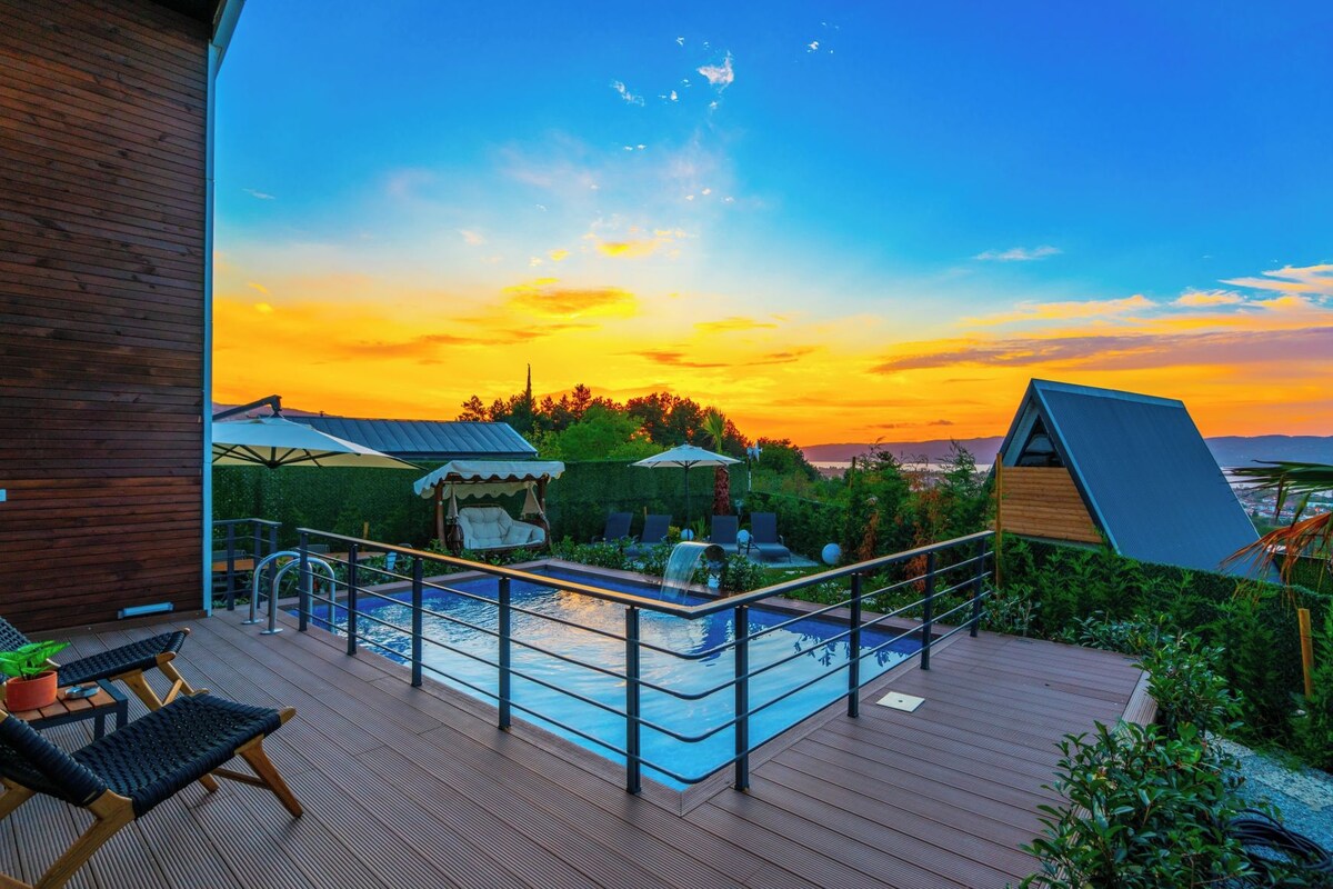 A pool area with a wooden deck, lounge chairs, and a view of the sunset over the ocean.