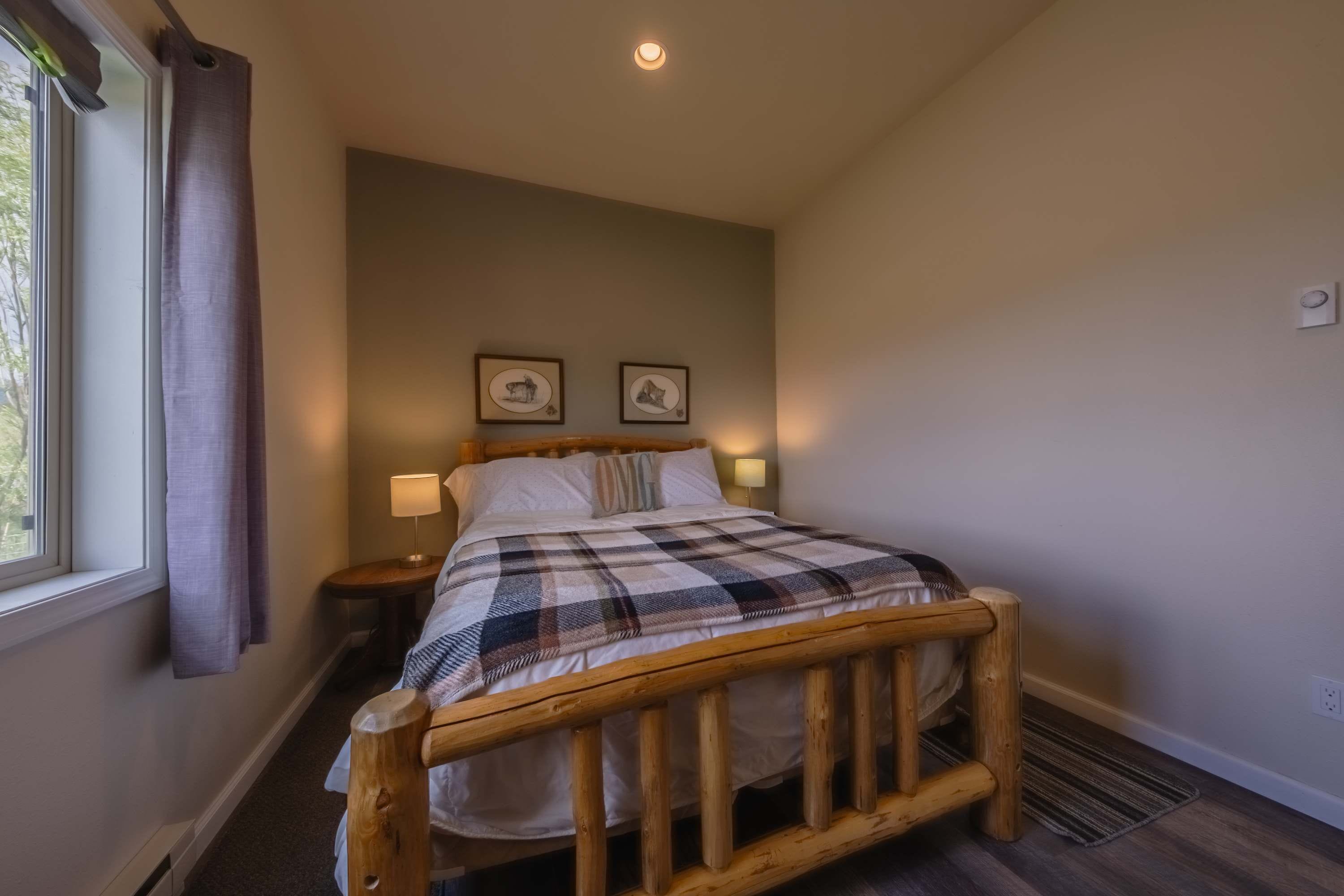 A bedroom with a wooden bed frame, a checkered blanket, two bedside lamps, and two framed pictures on the wall.