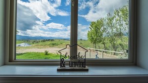 Une fenêtre donnant sur un champ verdoyant et des arbres, et un panneau en métal en forme de maison, sur lequel on peut lire les mots « respirez et détendez-vous », posé sur un rebord de fenêtre.