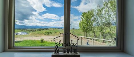 A window with a view of a grassy field and trees, a metal house-shaped sign with the words "breathe & unwind" on a windowsill.