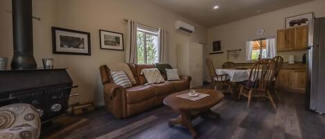 A cozy living room with a sofa, a coffee table, and a fireplace. There is a dining area with a table and chairs, and a kitchen with wooden cabinets and a refrigerator.