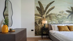 A bedroom with a bed, bedside table, lamp, and a wall mural of foliage.
