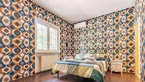 Une chambre à coucher avec un papier peint à motifs, une fenêtre, un radiateur, un lit, une table de chevet et une commode.