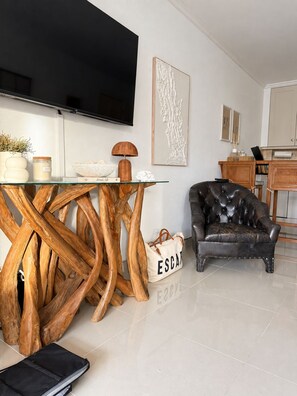 A modern living room with a glass-top coffee table, a leather armchair, a television mounted on the wall, and a dining area with wicker chairs.