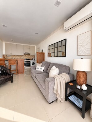 A modern living room with a grey sofa, a black armchair, a small black side table with a lamp, and a kitchen in the background. - Elegant Poolside Apartment, Large Covered Terrace- Walk to Beach, West Coast (Weston St James)