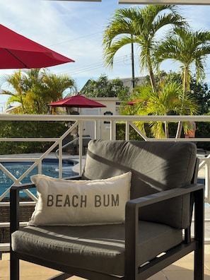 A lounge chair with a pillow labeled 'BEACH BUM' by a poolside, with palm trees and a red umbrella in the background.