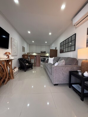 A modern living room with a grey sofa, a black armchair, a wooden coffee table, and a TV on the wall. - Elegant Poolside Apartment, Large Covered Terrace- Walk to Beach, West Coast (Weston St James)