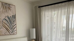 Un lit bien fait, avec du linge de lit blanc et un oreiller orné d’un motif, une table de chevet avec une lampe et une fenêtre avec des rideaux légers.