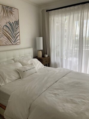A neatly made bed with white linens and a pillow with text, a bedside table with a lamp, and a window with sheer curtains. - Elegant Poolside Apartment, Large Covered Terrace- Walk to Beach, West Coast (Weston St James)
