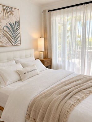 A bedroom with a bed, a bedside lamp, a nightstand, a window with sheer curtains, and a wall art piece. - Elegant Poolside Apartment, Large Covered Terrace- Walk to Beach, West Coast (Weston St James)