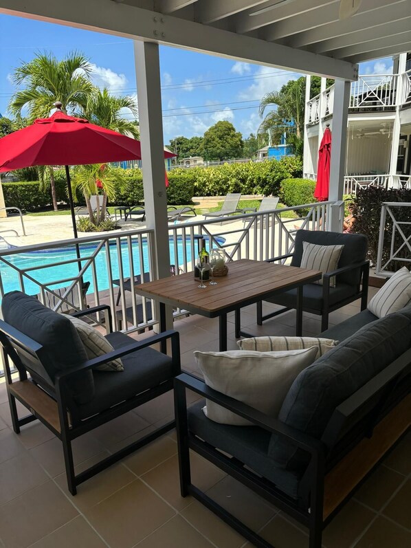 A covered outdoor seating area with a pool, lounge chairs, and a table with a red umbrella. - Elegant Poolside Apartment, Large Covered Terrace- Walk to Beach, West Coast (Weston St James)
