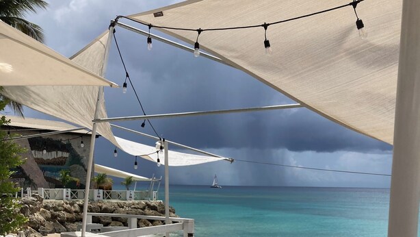 Une terrasse au bord de la plage, avec des tables et des chaises blanches, un panier en osier rempli de plantes, et une vue sur l’océan, où l’on aperçoit un voilier au loin.