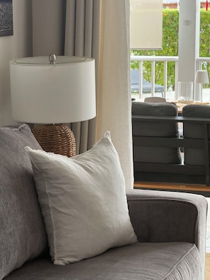 A grey sofa with a white pillow, a wicker lamp, and a view of an outdoor balcony with a table and chairs.