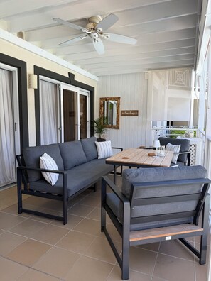 A covered patio area with a grey sofa, a wooden table, and a ceiling fan. - Elegant Poolside Apartment, Large Covered Terrace- Walk to Beach, West Coast (Weston St James)