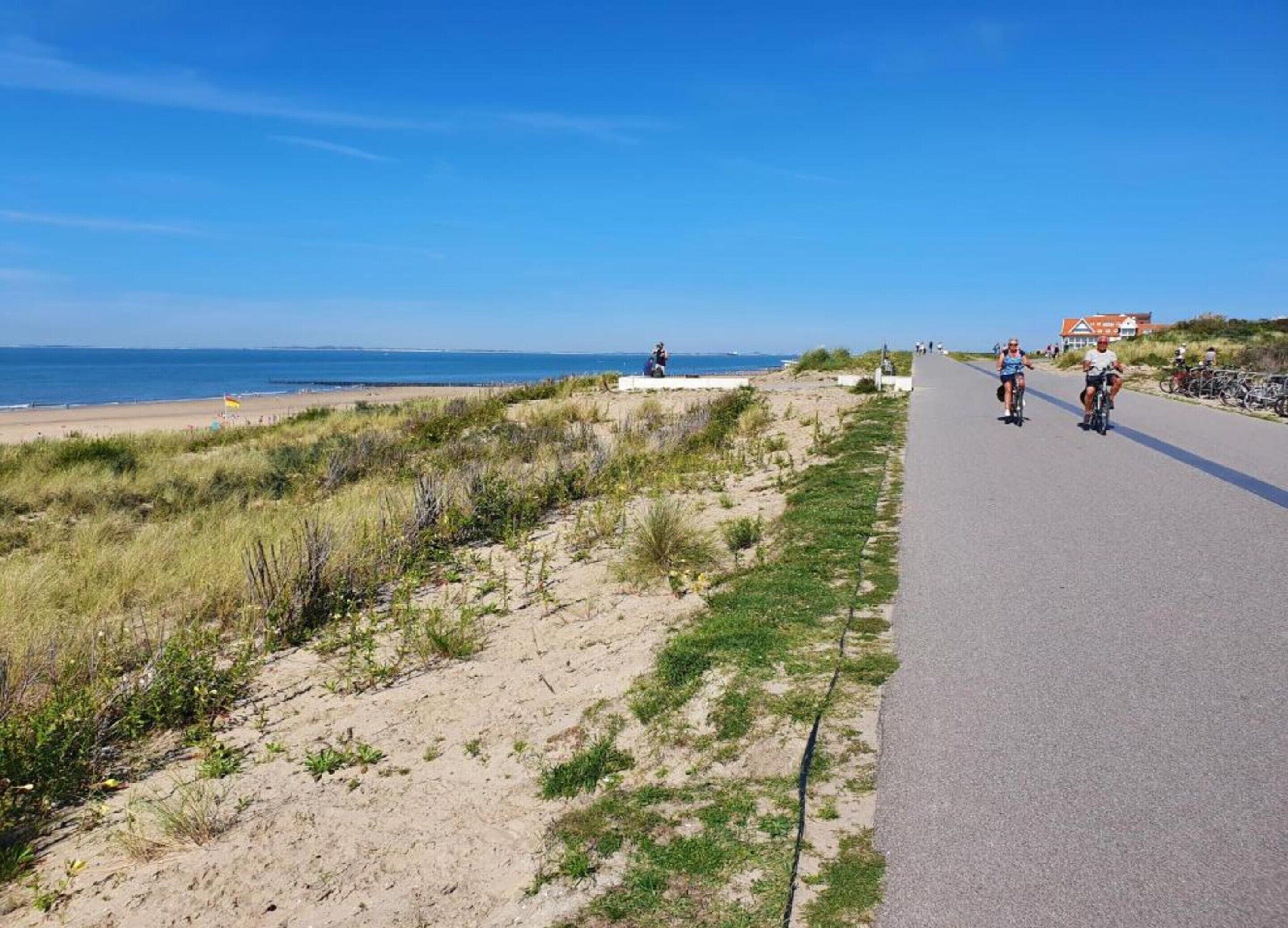 A coastal road with cyclists, a beach, and a building with a red roof in the background.