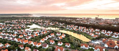 Vue aérienne d’un quartier résidentiel avec des maisons, une étendue d’eau et une silhouette de ville en arrière-plan, au coucher du soleil.
