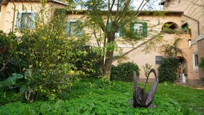 Ein Garten mit vielfältigen Pflanzen und einem Baum im Vordergrund, im Hintergrund ein Gebäude mit einer beige farbenen Fassade und grünen Fensterläden.