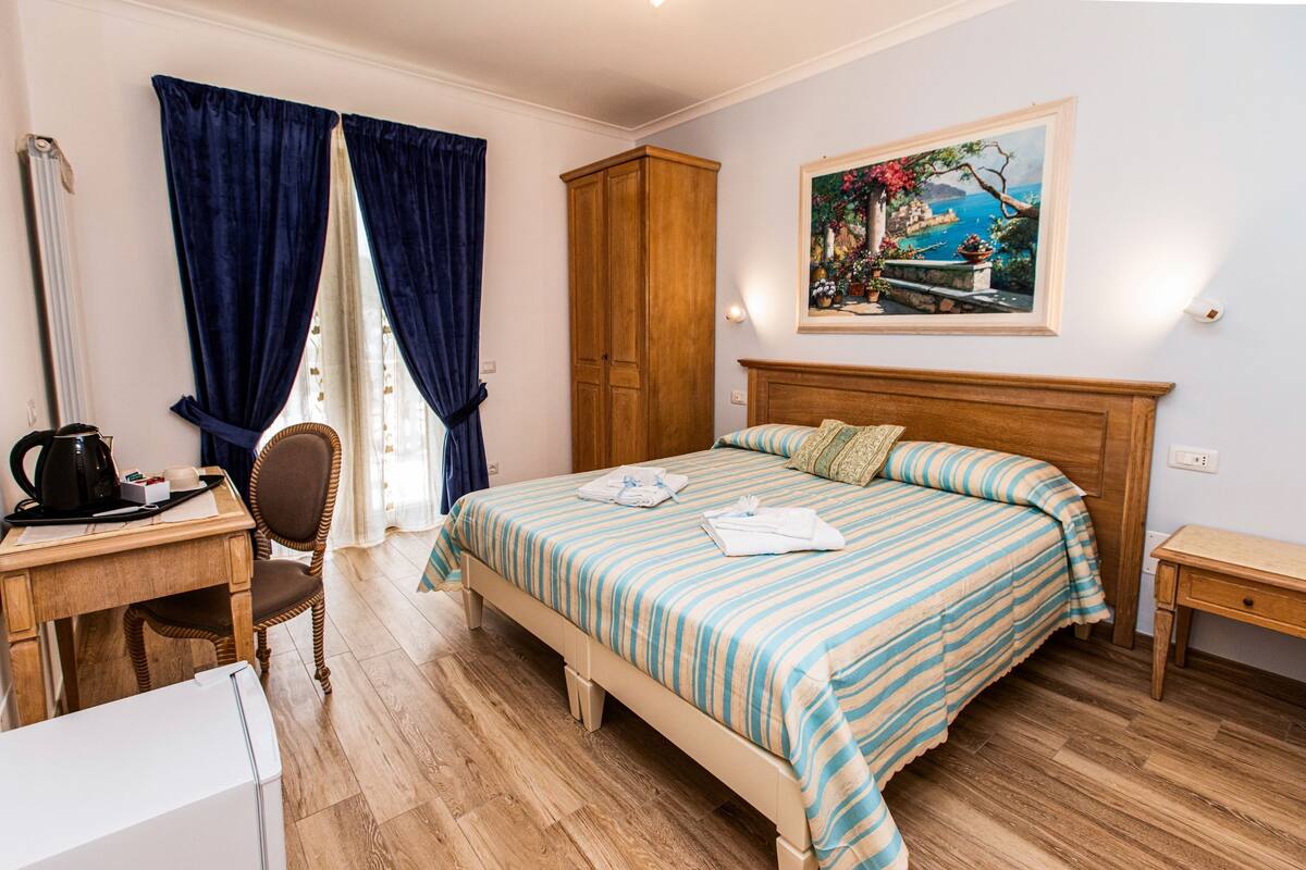 A bedroom with a bed, a wooden wardrobe, a desk with a kettle, and a chair.