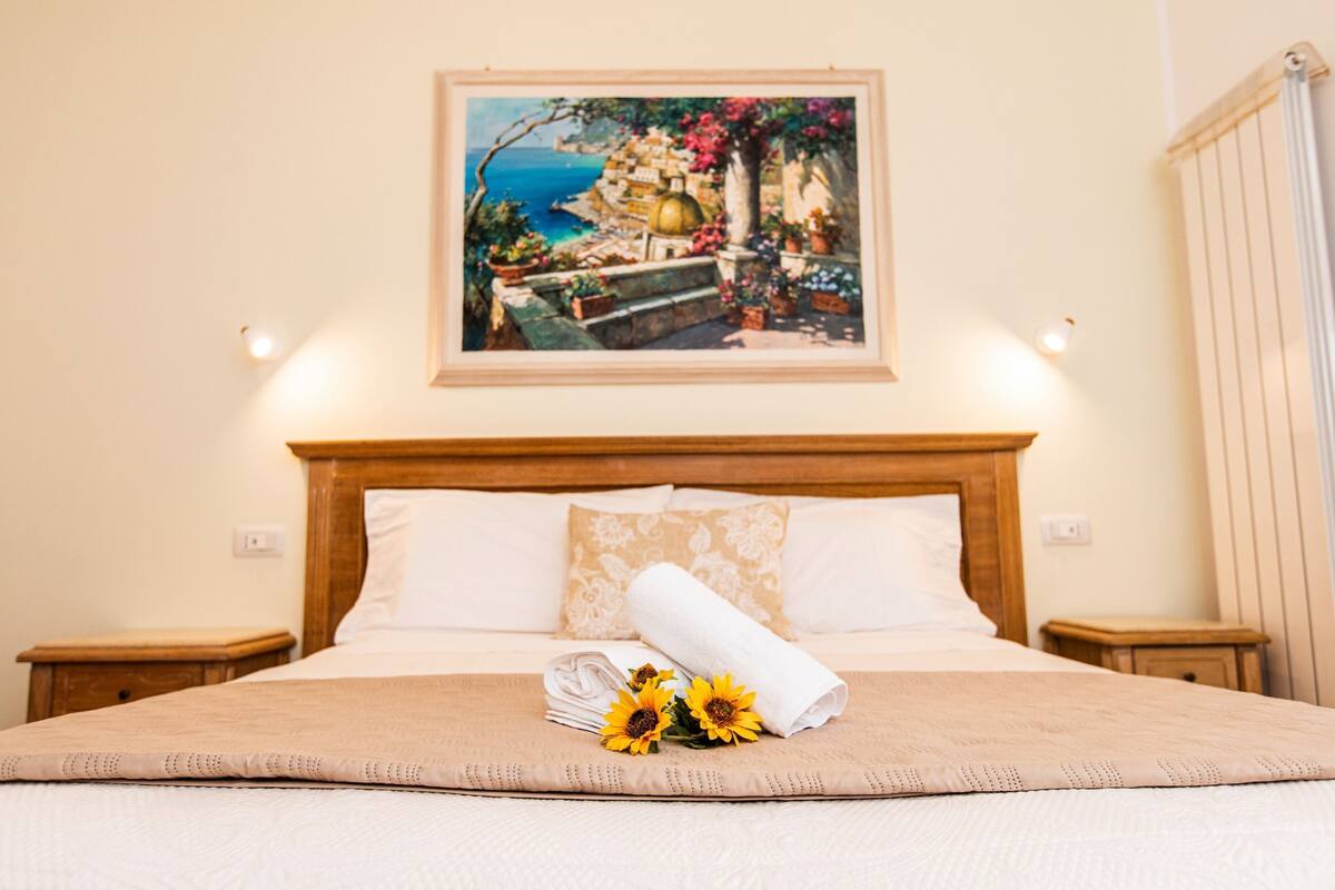 A neatly made bed with a beige bedspread, a rolled white towel, and two sunflowers. A framed painting of a coastal scene hangs above the bed.