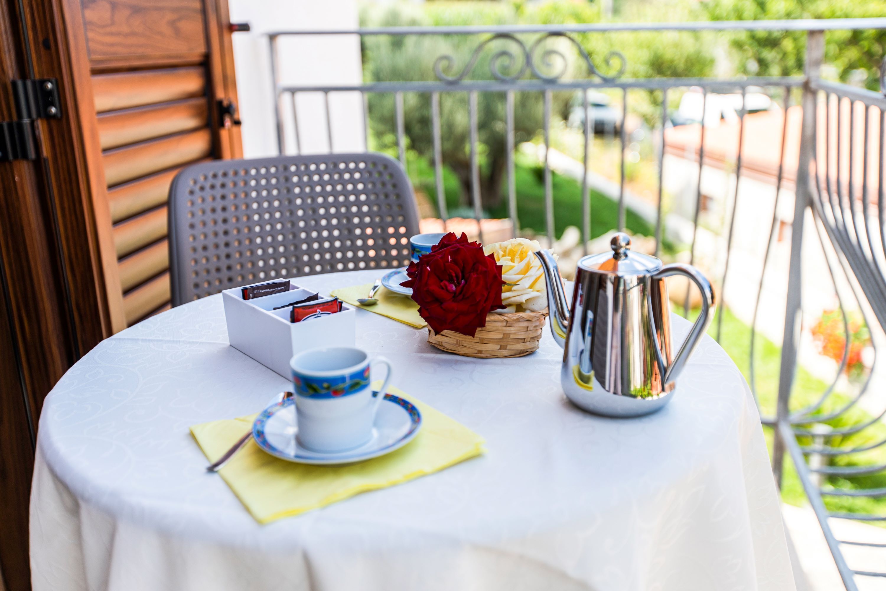 Ein runder Tisch mit weißer Tischdecke, einer blau-weißen Tasse, einer roten Rose, einem Korb und einem silbernen Teekessel auf einem Balkon mit Metallgeländer.
