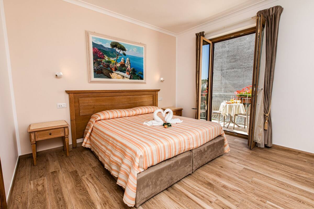 A bedroom with a wooden bed, a nightstand, a painting of a coastal scene, and a balcony with a table and chairs.