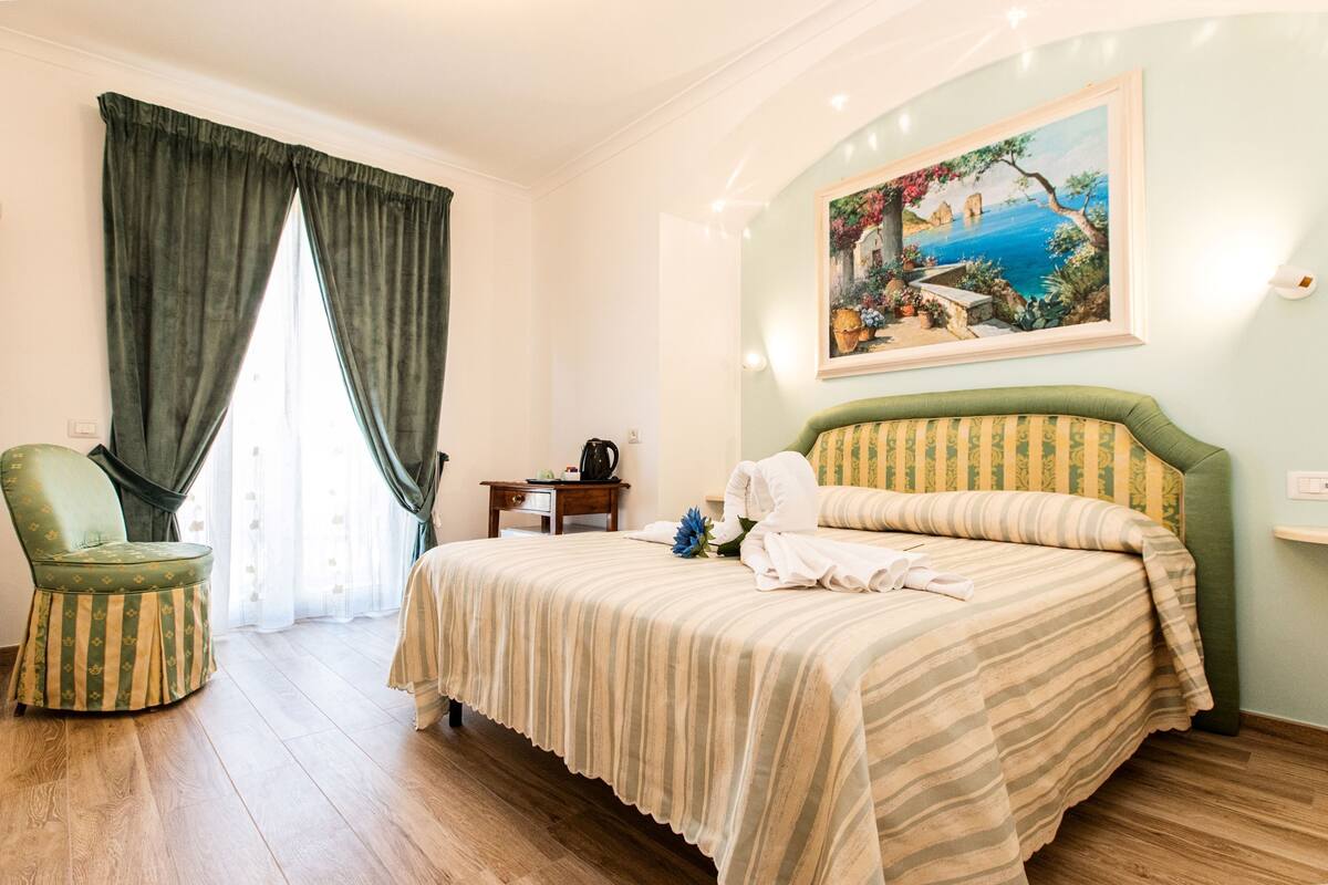 A hotel room with a bed, a chair, a desk, and a painting on the wall.