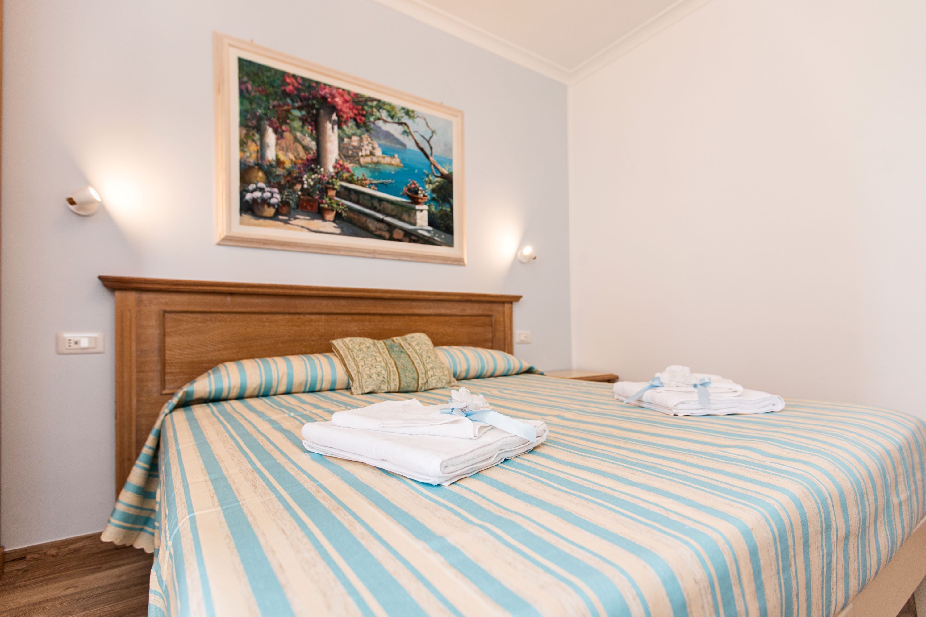 A neatly made bed with striped bedding and pillows, a framed painting above it, and a wooden headboard.