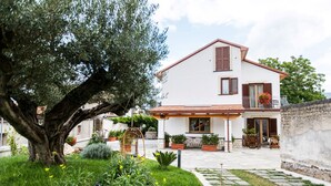 A white two-story house with a tiled roof, a balcony with wooden shutters, and a garden with a large tree and potted plants. - Al Chiaro di Luna (Agerola)