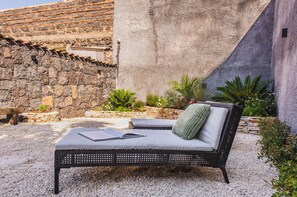 A wicker sofa with cushions and a book, set against a stone wall and surrounded by plants. - Leone Apartment with Garden by Wonderful Italy (Catania)