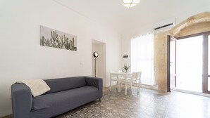 Un salon avec un canapé gris, un sol à motifs, un tableau représentant un cactus, une table avec des chaises et une porte donnant sur l’extérieur.