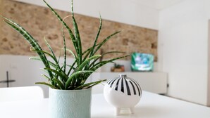 Une plante en pot avec de longues feuilles fines, placée dans un pot à texture, un petit vase blanc à rayures noires et un téléviseur en arrière-plan.