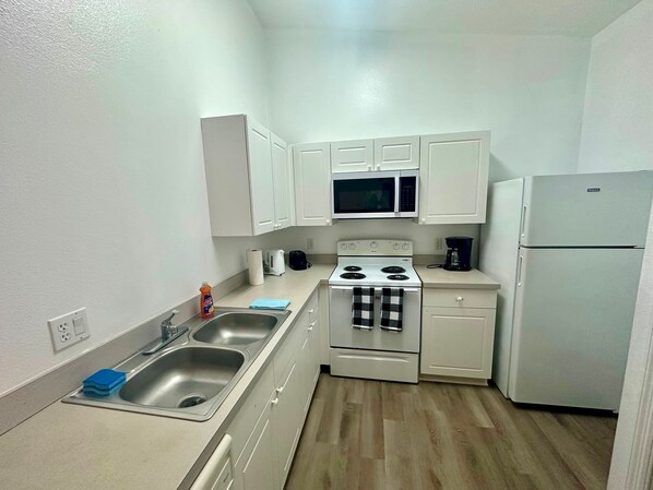 Una cocina compacta con armarios blancos, un lavamanos de acero inoxidable, un microondas, una estufa y un refrigerador.