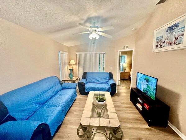 Un salon avec un ventilateur de plafond, un téléviseur, un canapé, une table basse et une lampe.