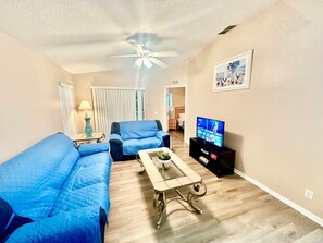 Un salon avec un ventilateur de plafond, un téléviseur, un canapé, un fauteuil et une table basse.