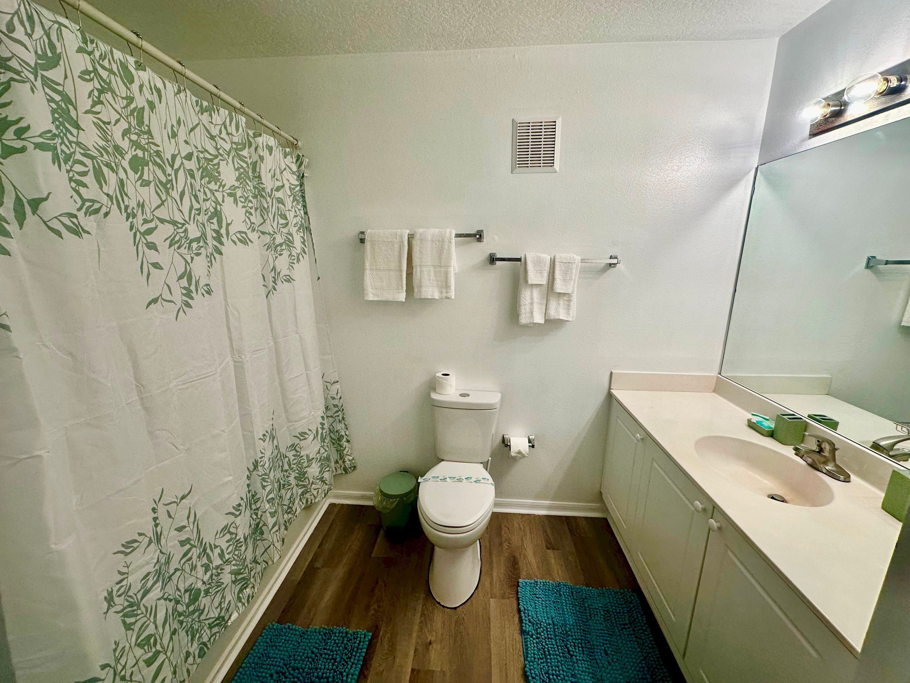 A bathroom with a toilet, sink, and mirror. There are towels hanging on a rack and a shower curtain with a leaf pattern.