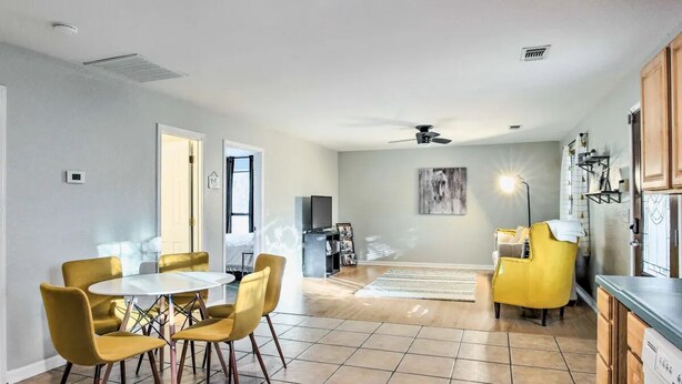 A modern kitchen with wooden cabinets, a dining area with a round table and yellow chairs, and a living room with a yellow armchair and a painting on the wall.